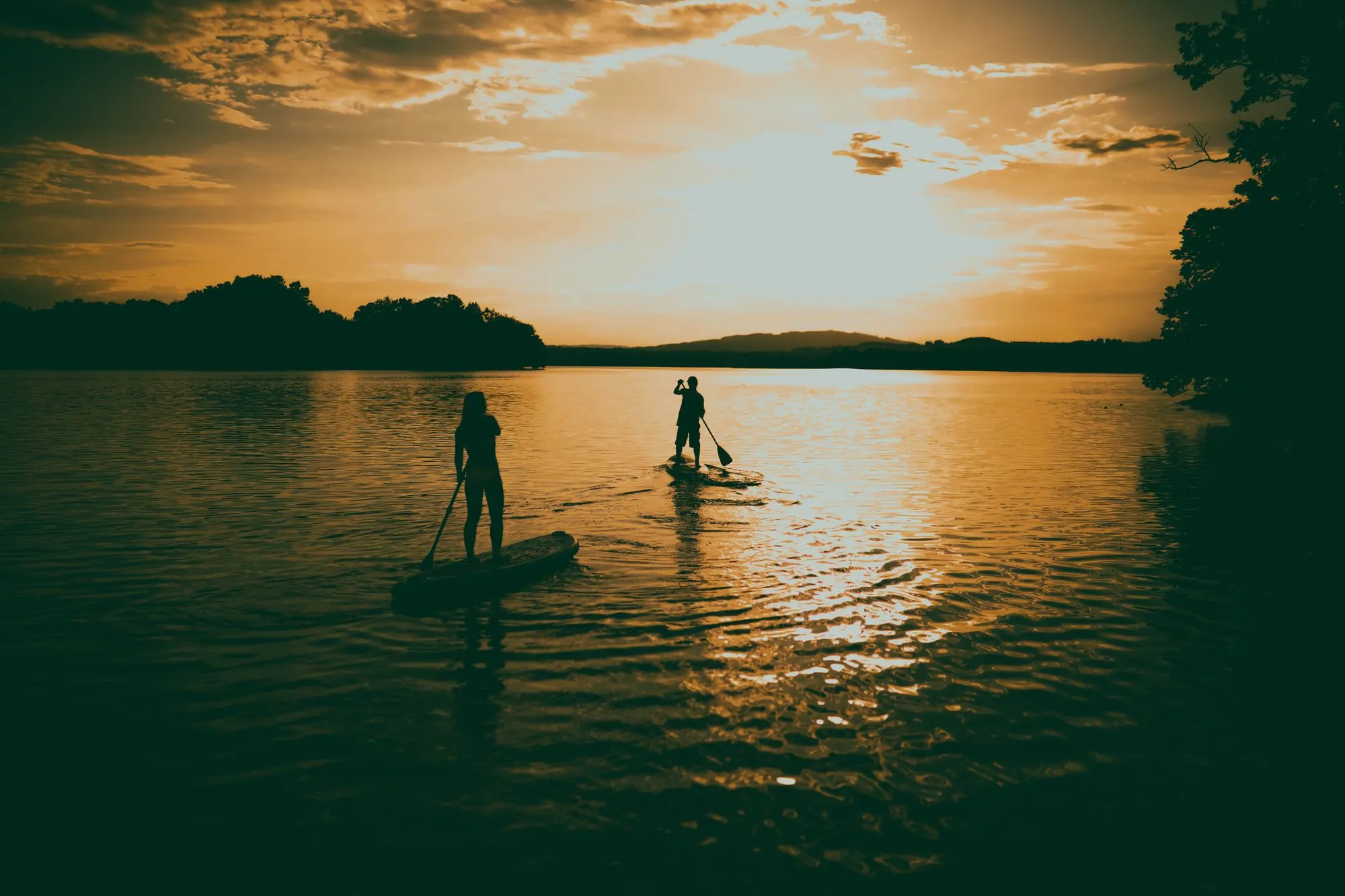 Two paddleboarders standing on their boards on a calm lake at sunset, with trees on the shore
